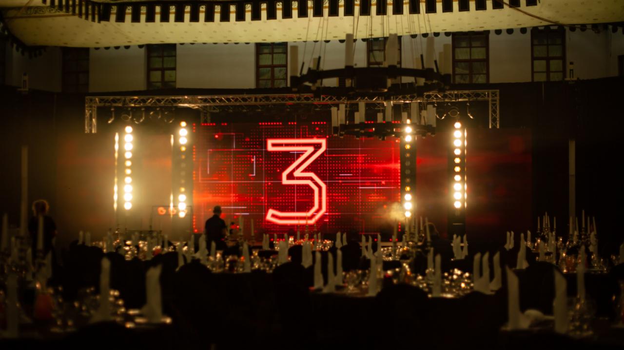 Sala bankietowa przygotowana na uroczystość, scena z ekranem LED wyświetlającym cyfrę 3, oświetlenie punktowe, nakryte stoły w tle.