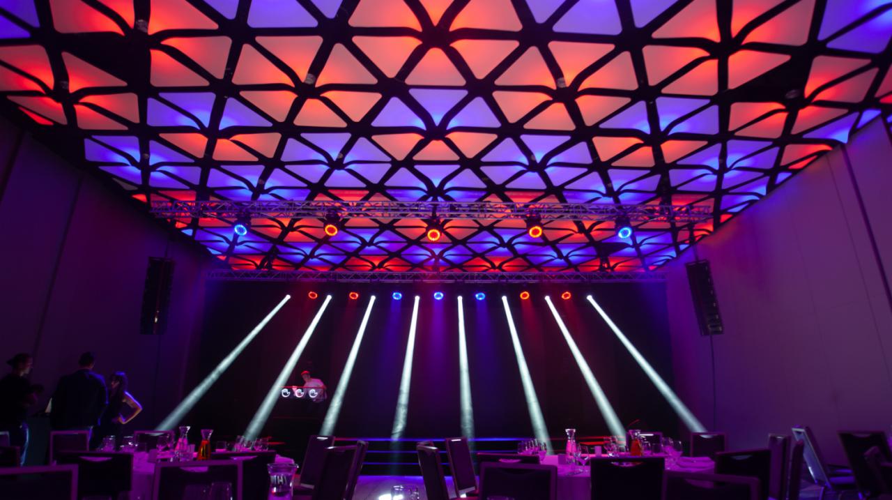 Sala bankietowa z geometrycznym sufitem podświetlonym na czerwono i niebiesko, reflektory sceniczne oświetlają DJ-a za konsoletą, przygotowane stoły dla gości.