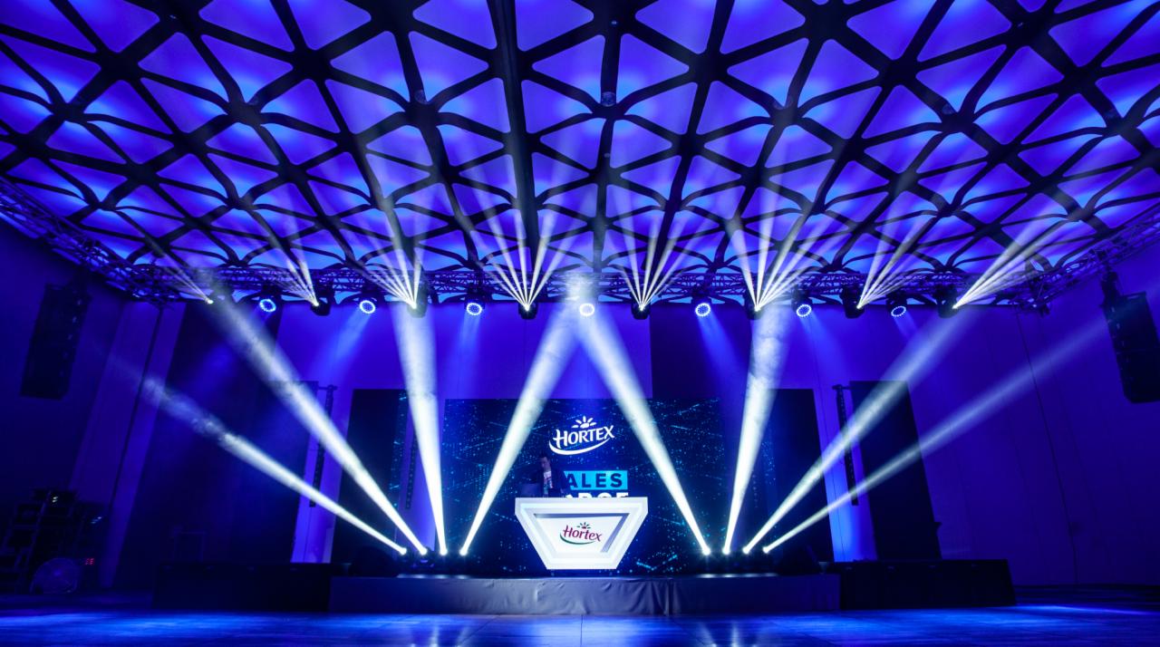 Scena oświetlona niebieskimi światłami, geometryczny sufit, logo Hortex na froncie podświetlonego pulpitu, osoba w tle za pulpitem.