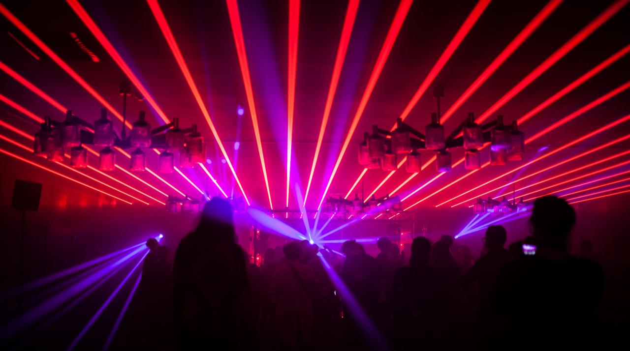 Wnętrze klubu oświetlone czerwonymi i fioletowymi laserami, z sylwetkami ludzi w tle i ozdobnym żyrandolem na suficie.