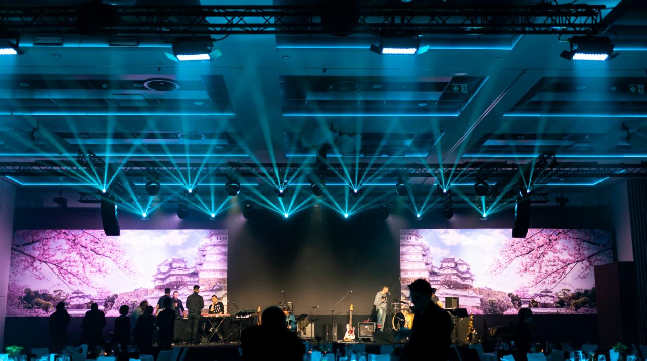 Scena z zespołem muzycznym podświetlona niebieskim oświetleniem scenicznym, z wyświetlanym obrazem kwitnącej wiśni i japońskiego zamku w tle.