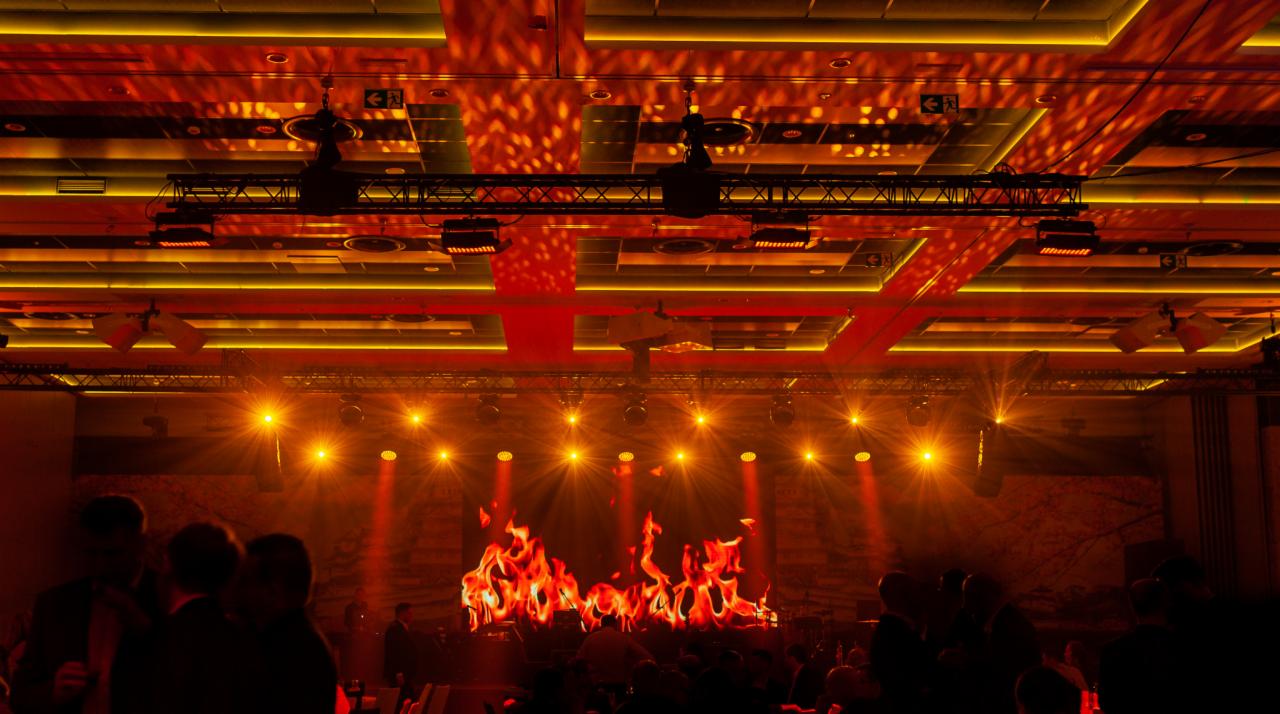 Scena oświetlona pomarańczowymi reflektorami, z ekranem LED wyświetlającym animację ognia i sylwetkami publiczności w tle, w sali z dekoracyjnym oświetleniem sufitowym.