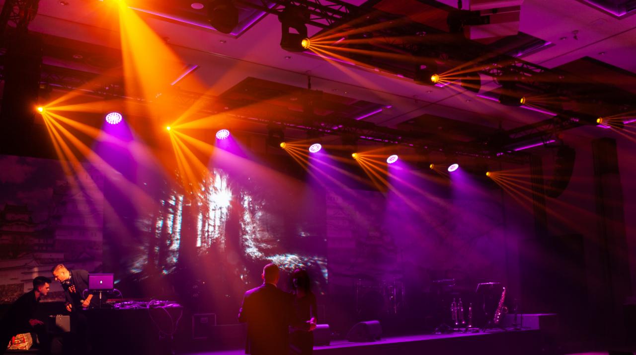 Scena oświetlona pomarańczowymi i fioletowymi reflektorami, z parą tańczącą na tle wyświetlanego lasu i pracującym DJ-em z laptopem.