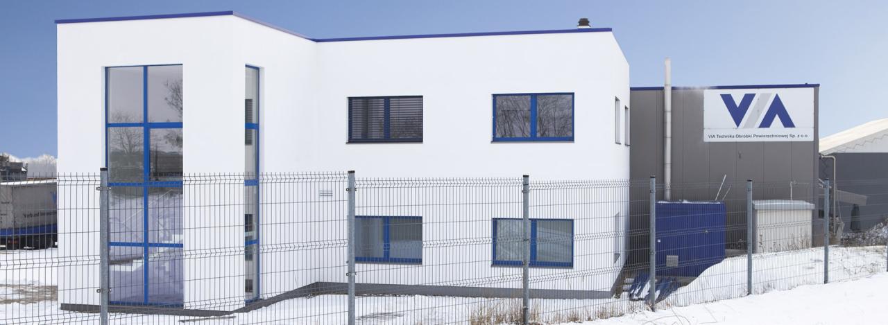 Nowoczesny budynek firmy VA Technika Obróbki Powierzchniowej Sp. z o.o. z białymi ścianami, niebieskimi ramami okiennymi i stalową konstrukcją ogrodzenia na ośnieżonym terenie.