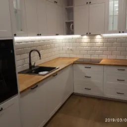 Nowoczesna, biała kuchnia z drewnianym blatem, czarnym zlewem i baterią, oraz białymi płytkami metra na ścianie, oświetlona listwą LED pod szafkami wiszącymi.