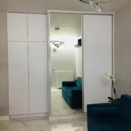 Biała szafa przesuwna z lustrem, widoczny fragment turkusowej sofy, marmurowa podłoga i nowoczesny żyrandol.