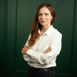 Portret kobiety z rudymi włosami w białej koszuli i czarnych spodniach, stojącej na tle ciemnozielonej ściany z panelami.