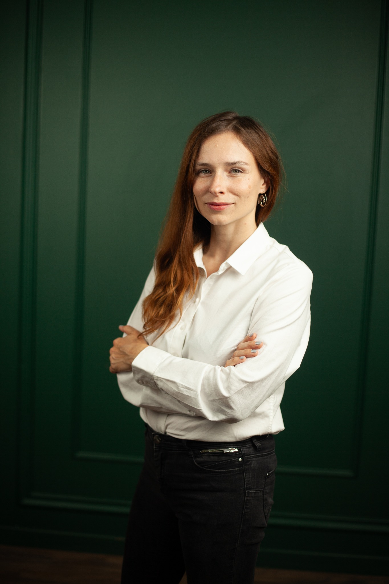 Portret kobiety z rudymi włosami w białej koszuli i czarnych spodniach, stojącej na tle ciemnozielonej ściany z panelami.