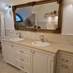 Biała szafka łazienkowa z dwoma umywalkami, beżowym blatem i bogato zdobionym, złotym lustrem w stylu retro.