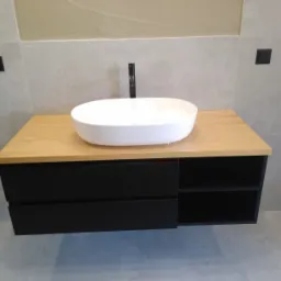 Biała, owalna umywalka nablatowa na blacie z jasnego drewna, czarne szafki z szufladami i otwartymi półkami, szare płytki na ścianie i podłodze, minimalistyczna armatura.