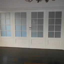 Czteroskrzydłowe, białe drzwi wewnętrzne z przeszkleniami, widoczne z niskiej perspektywy na tle ciemnej podłogi i jasnych ścian.