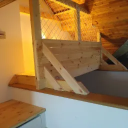 Drewniana konstrukcja antresoli z barierką z siatki sznurkowej, widok z dołu na drewniany sufit ze skosu.