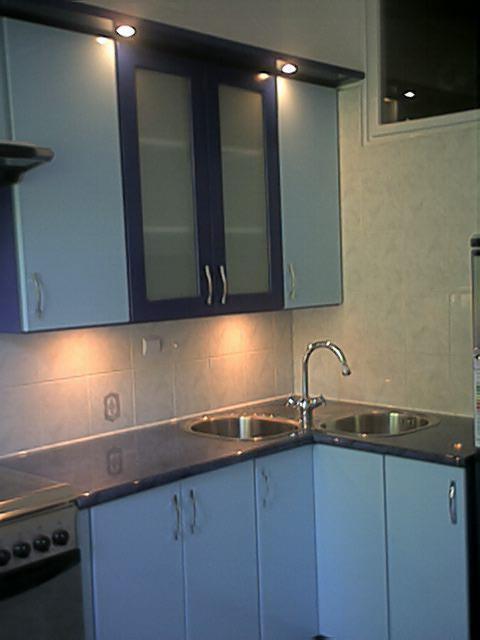 Narożna zabudowa kuchenna z dwukomorowym zlewem, jasnoniebieskimi frontami szafek i ciemnogranatowymi ramami przeszklonych szafek wiszących, oświetlona punktowymi lampkami podszafkowymi.