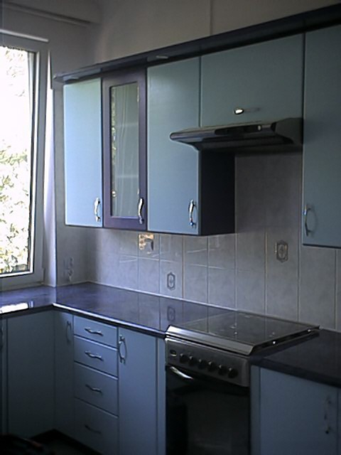 Niebieska zabudowa kuchenna z ciemnymi blatami i szarą okapem, widok na okno z firanką, płytki ceramiczne na ścianie.