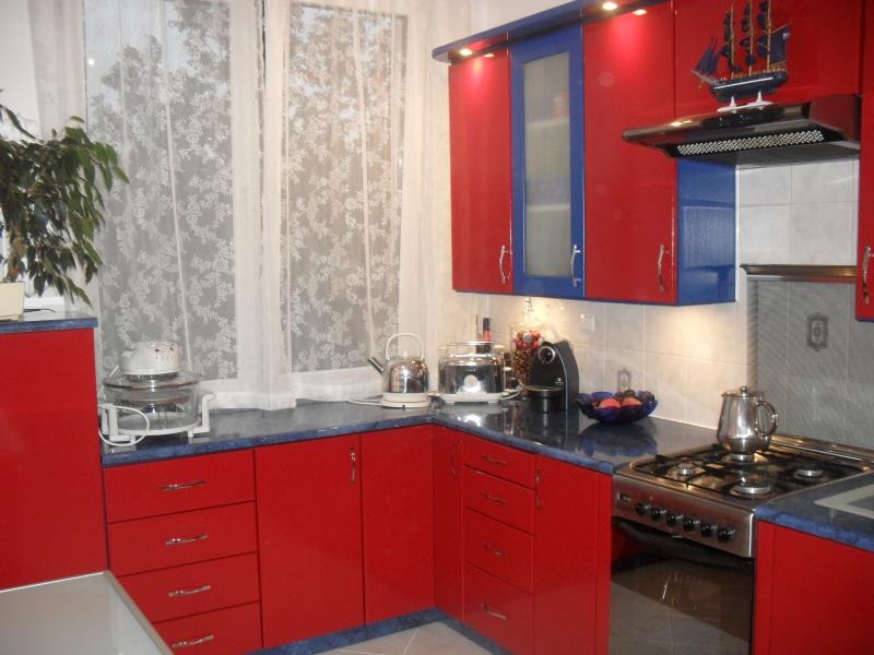 Czerwono-niebieska kuchnia z ciemnym blatem, widoczny fragment okna z koronkową firanką, na blacie czajnik, toster i ekspres do kawy.