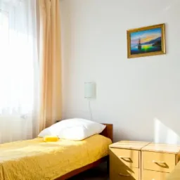 Pokój dwuosobowy z dwoma pojedynczymi łóżkami przykrytymi żółtymi narzutami, białymi poduszkami i żółtymi ręcznikami, jasne zasłony w oknie i obraz z motywem morskim na ścianie.