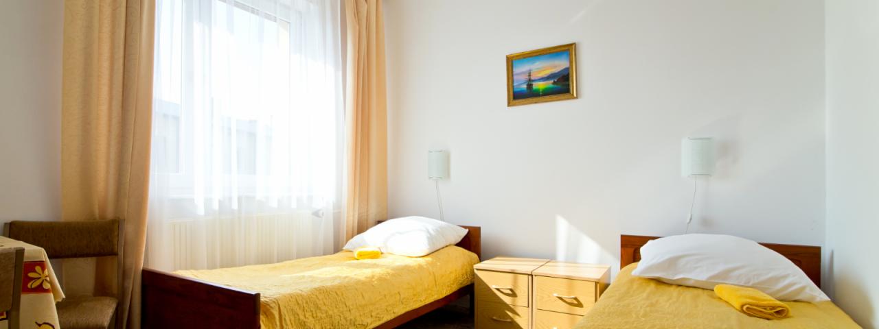 Pokój dwuosobowy z dwoma pojedynczymi łóżkami przykrytymi żółtymi narzutami, białymi poduszkami i żółtymi ręcznikami, jasne zasłony w oknie i obraz z motywem morskim na ścianie.