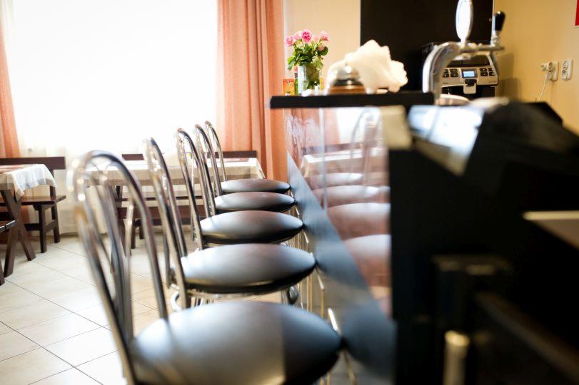 Rząd chromowanych krzeseł barowych z czarnymi siedziskami ustawiony przy błyszczącym, czarnym barze; w tle okno z jasnoróżowymi zasłonami i wazon z różowymi różami.