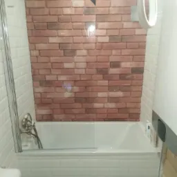 Biała wanna obudowana białymi kafelkami typu metro, z parawanem nawannowym i baterią, na ścianie dekoracyjna tapeta imitująca czerwoną cegłę.