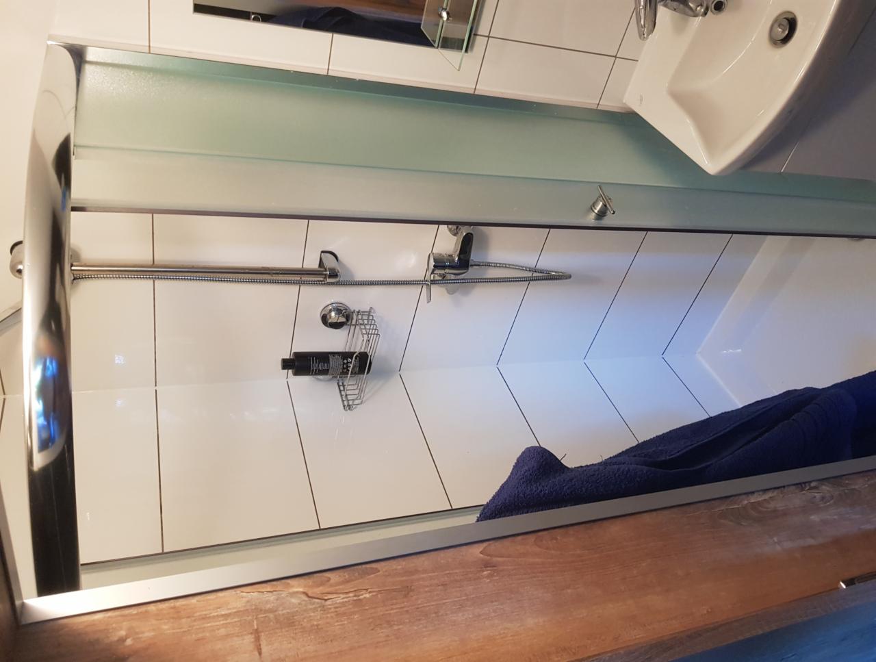 Kabina prysznicowa z białymi płytkami ułożonymi w jodełkę, chromowaną armaturą i półką na kosmetyki, widoczny fragment umywalki i niebieski ręcznik.