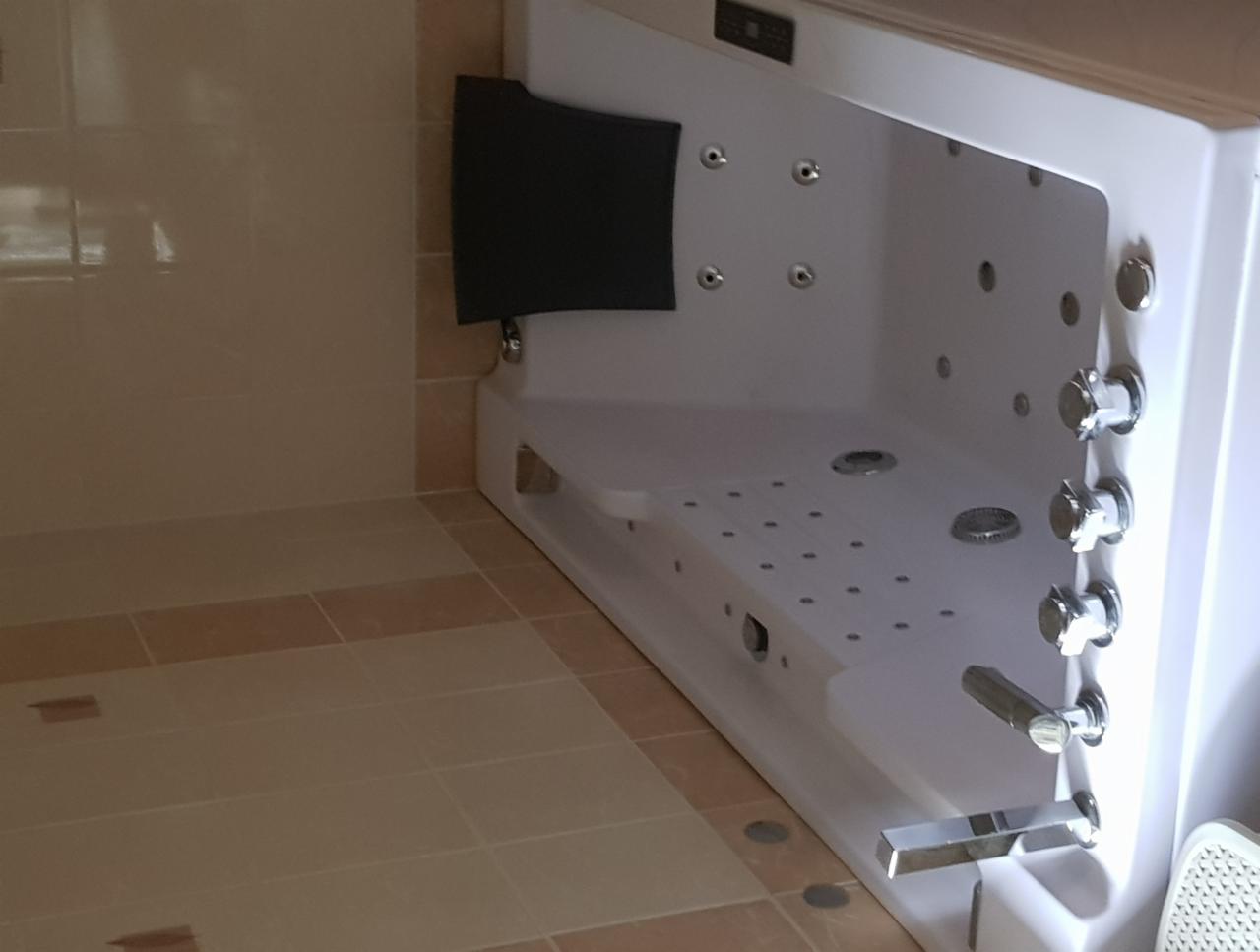 Nowoczesna wanna z hydromasażem w jasnej łazience, widoczne dysze, chromowane baterie i panel sterowania, płytki podłogowe w odcieniach beżu i brązu.