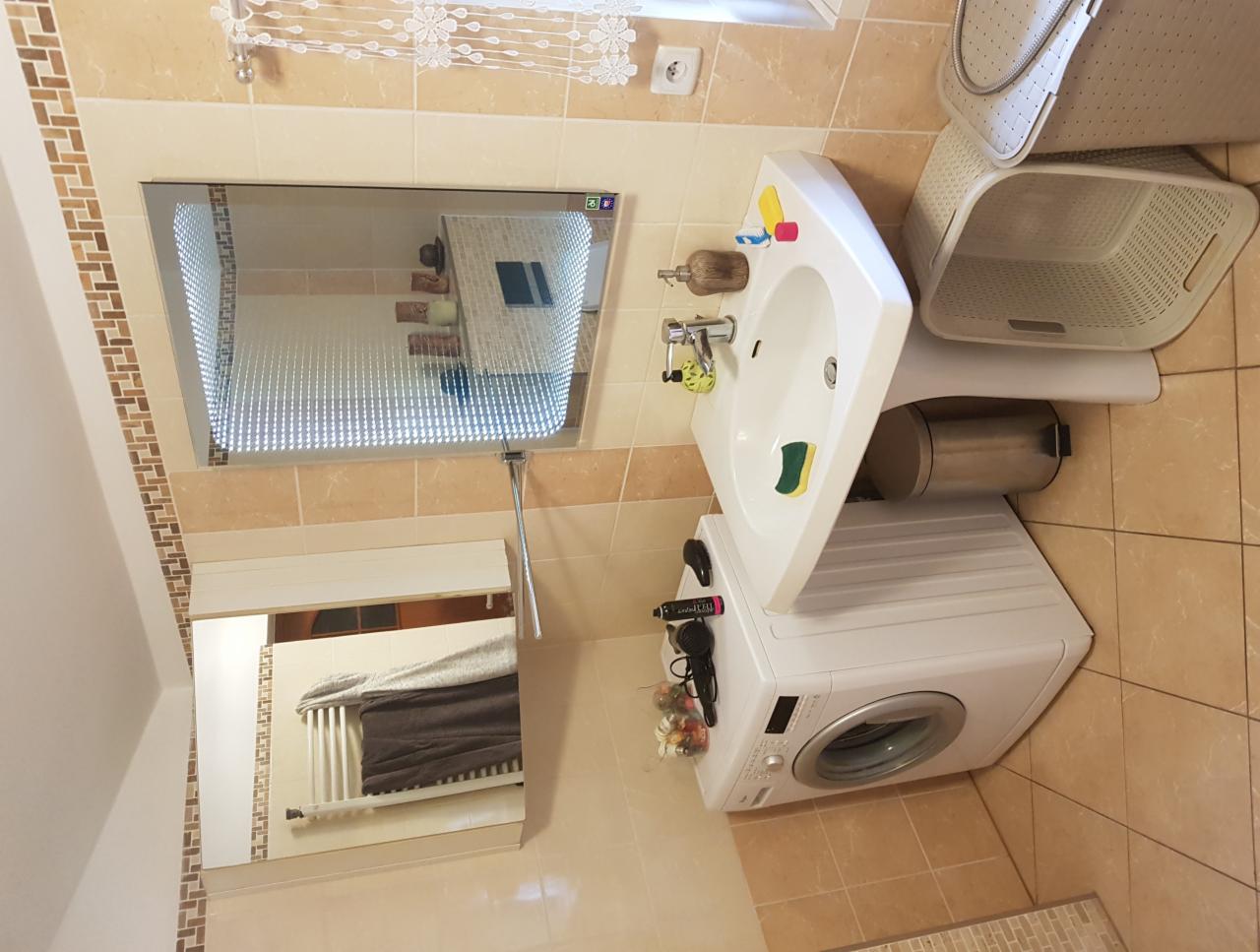 Jasna łazienka z beżowymi płytkami, umywalką na postumencie, pralką, koszem na pranie i lustrami z oświetleniem LED, odbijającymi wnętrze pomieszczenia.