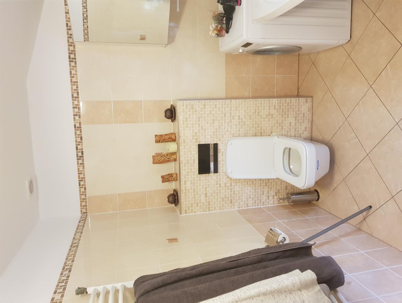 Beżowa łazienka z podwieszaną toaletą, mozaiką na ścianie, pralką i dekoracyjnymi elementami.