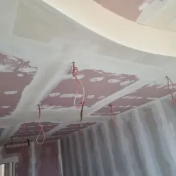 Ściany i sufit w trakcie szpachlowania, widoczne kable elektryczne zwisające z sufitu, płyty gipsowo-kartonowe na ścianach i suficie, częściowo pokryte białą masą szpachlową.