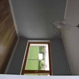 Usługi Budowlane Łukasz Jaromieńczuk - Pomieszczenie po malowaniu ścian na szaro, z widocznym oknem, podłogą z paneli i lampą zabezpieczoną folią.