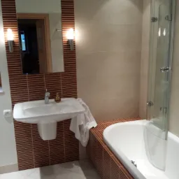 Umywalka wisząca z lustrem i oświetleniem, obok wanna z parawanem, wykończone brązowymi płytkami w poziomie i beżowymi płytkami na ścianach