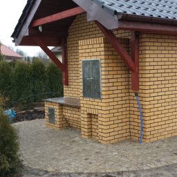 Usługi Budowlane Łukasz Jaromieńczuk - Murowana altana z cegły klinkierowej z ciemnoszarą dachówką, drewnianymi elementami konstrukcyjnymi w kolorze wiśniowym, metalowymi drzwiczkami i niebieskim wężem ogrodowym, otoczona brukowaną...