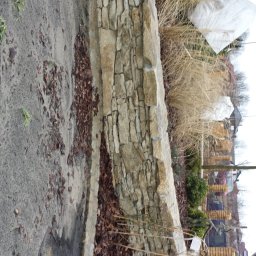 Usługi Budowlane Łukasz Jaromieńczuk - Kamienny murek oporowy z piaskowca, oddzielający ścieżkę od rabaty z trawami i krzewami, widok z góry na tle zabudowań.