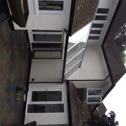 Usługi Budowlane Łukasz Jaromieńczuk - Elewacja domu z białego ceglanego muru z białymi oknami i brązowym dachem, widok z poziomu patio wyłożonego ciemną, błyszczącą kostką.