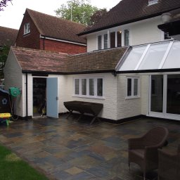 Usługi Budowlane Łukasz Jaromieńczuk - Wyłożony kamienną płytą taras z dwoma wiklinowymi fotelami, przylegający do białego budynku z przeszklonym dachem i otwartymi drzwiami do garażu z zielonym wężem ogrodowym na ścianie.