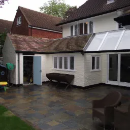 Wyłożony kamienną płytą taras z dwoma wiklinowymi fotelami, przylegający do białego budynku z przeszklonym dachem i otwartymi drzwiami do garażu z zielonym wężem ogrodowym na ścianie.