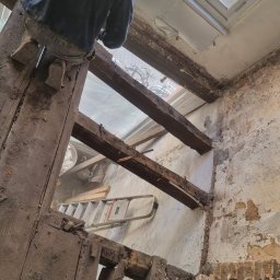 Usługi Budowlane Łukasz Jaromieńczuk - Pracownik na drabinie podczas demontażu drewnianej konstrukcji stropu w pomieszczeniu w trakcie generalnego remontu, widoczne odsłonięte cegły i fragmenty starych tynków.