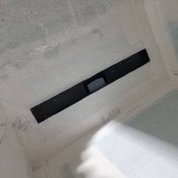 Usługi Budowlane Łukasz Jaromieńczuk - Niedokończona łazienka z odpływem liniowym w podłodze, ściany przygotowane pod wykończenie, widoczne ślady szpachlowania na ścianach i podłodze, zabezpieczone folią.