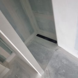 Usługi Budowlane Łukasz Jaromieńczuk - Przygotowanie do wylewki w narożniku łazienki z zamontowanym odpływem liniowym, widoczne ściany z płyt gipsowo-kartonowych i białe profile wykończeniowe.