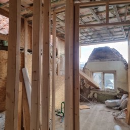 Usługi Budowlane Łukasz Jaromieńczuk - Wnętrze w trakcie przebudowy: widoczne drewniane słupki konstrukcyjne, płyty OSB na ścianach, ocieplenie dachu z folią, a w tle fragment zrujnowanej ściany z oknem.