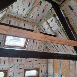 Usługi Budowlane Łukasz Jaromieńczuk - Ocieplenie poddasza wełną mineralną i pianką montażową, widoczne drewniane belki konstrukcyjne i okna dachowe, folia izolacyjna z nadrukiem producenta.