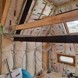 Usługi Budowlane Łukasz Jaromieńczuk - Poddasze w trakcie izolacji: widoczne krokwie, wełna mineralna, folia termoizolacyjna z nadrukiem, elementy konstrukcji stalowej, okno dachowe, przewody elektryczne i narzędzia na podłodze z płyty...