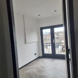 Wnętrze pokoju z parkietem ułożonym w jodełkę, widok przez otwarte drzwi na balkon z czarną balustradą i widokiem na zabudowę miejską.