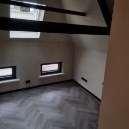 Poddasze po remoncie z ułożonymi panelami podłogowymi w jodełkę, widoczne okna połaciowe i wykończone ściany w jasnym odcieniu.