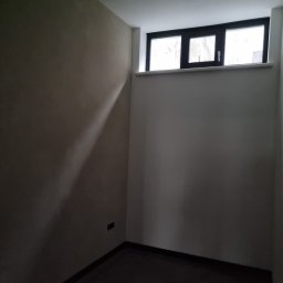 Usługi Budowlane Łukasz Jaromieńczuk - Wnętrze pokoju po szpachlowaniu i malowaniu ścian na beżowo, z ciemną podłogą, oświetleniem sufitowym i oknem z czarną ramą