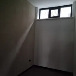 Wnętrze pokoju po szpachlowaniu i malowaniu ścian na beżowo, z ciemną podłogą, oświetleniem sufitowym i oknem z czarną ramą