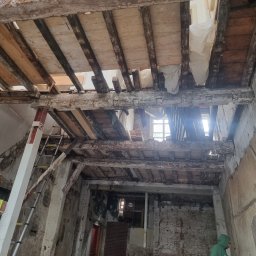 Usługi Budowlane Łukasz Jaromieńczuk - Wnętrze w trakcie demontażu starego stropu z drewnianymi belkami, widoczne fragmenty cegieł, instalacji i drabina oparta o filar, pracownik w zielonej bluzie przy ścianie.