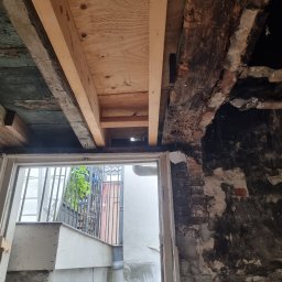 Usługi Budowlane Łukasz Jaromieńczuk - Fragment remontowanego pomieszczenia z odsłoniętym drewnianym stropem, fragmentem muru z cegły i widokiem na zewnątrz przez drzwi.