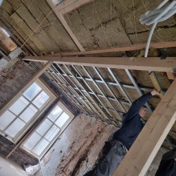 Usługi Budowlane Łukasz Jaromieńczuk - Montaż sufitu podwieszanego z wełną mineralną w pomieszczeniu z oknami, widoczny pracownik z poziomicą i fragmenty ściany z odsłoniętą cegłą.