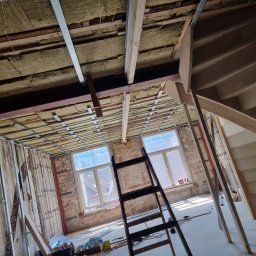 Usługi Budowlane Łukasz Jaromieńczuk - Wnętrze w trakcie prac: izolacja stropu wełną mineralną, metalowy stelaż pod sufitem, surowe ściany z cegły, drewniana konstrukcja schodów, drabina oparta o ścianę.