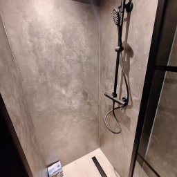 Usługi Budowlane Łukasz Jaromieńczuk - Nowoczesna kabina prysznicowa z szarą, teksturowaną ścianą, czarną armaturą i odpływem liniowym, z widocznym opakowaniem po produkcie w rogu.